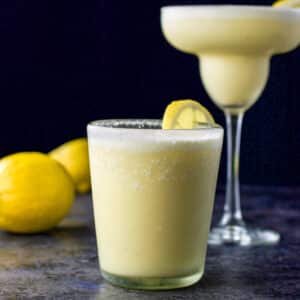Double old fashioned glass filled with the margarita in front with the classic margarita glass in the background. There are lemons on the board