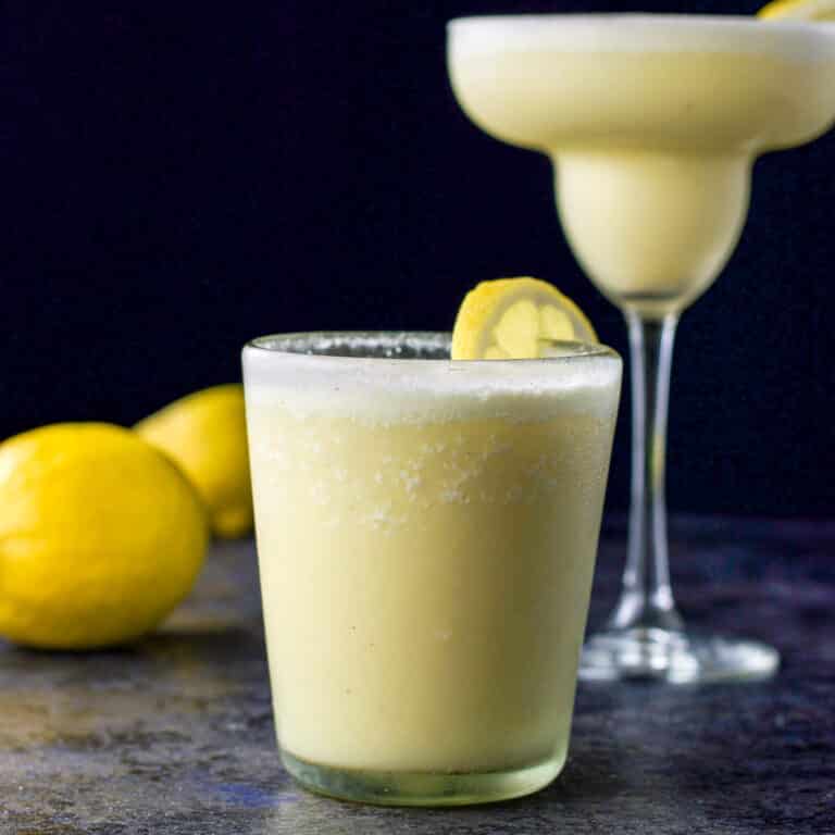 Double old fashioned glass filled with the margarita in front with the classic margarita glass in the background. There are lemons on the board