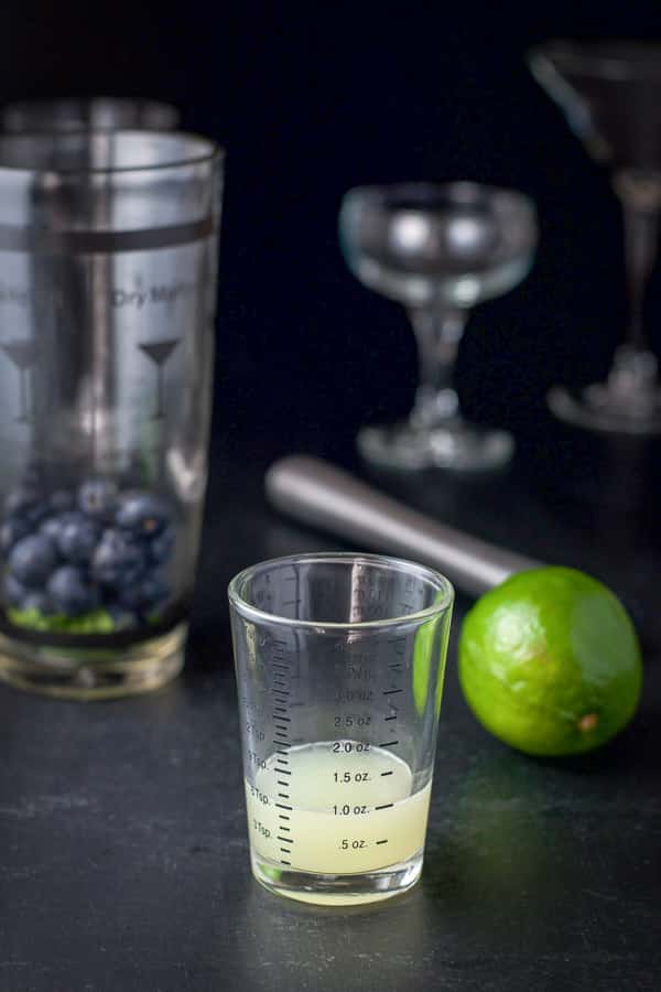 Lime juice measured out with the shaker in the background along with glasses, a lime and the muddler