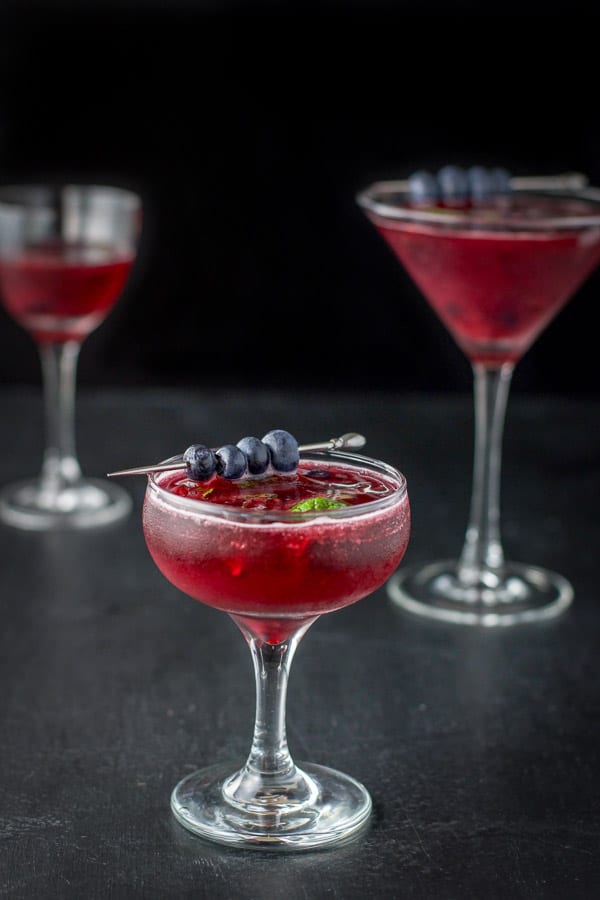 Blueberry cosmo with blueberries and mint leaves for garnish in a coupe glass in front and a martini glass in back