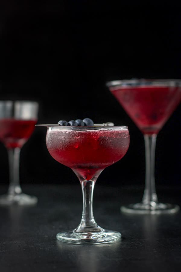 Vertical view of the blueberry cosmo with blueberries and mint leaves for garnish in a coupe glass in front and a martini glass in back