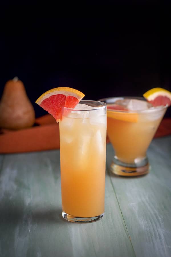 Tall Collins glass in front of the martini glass filled with the orange cocktail. There are grapefruit wedges on the side of the glasses