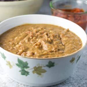A bowl filled with creamy pinto refried beans with some salsa in the background - square