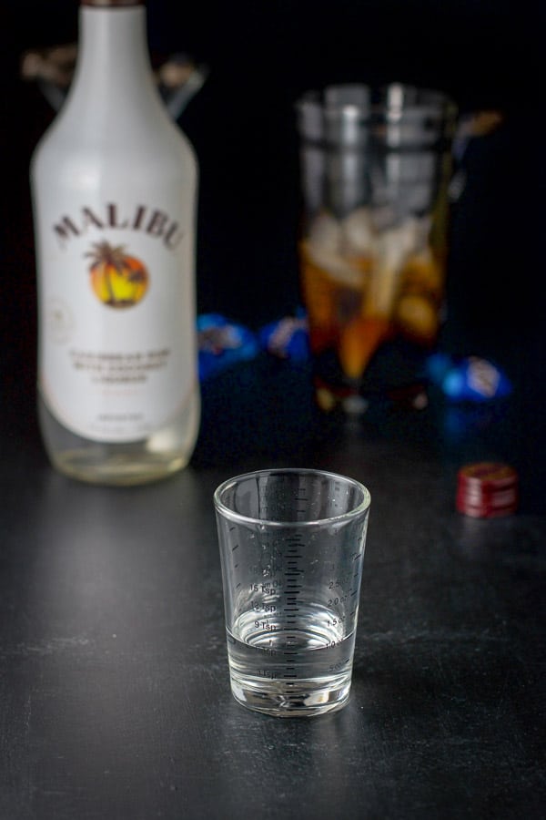 Coconut rum measured out with the bottle, shaker, candy bars and glasses in the background
