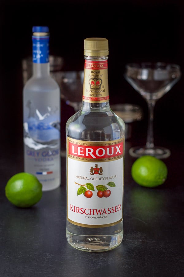 Cherry brandy, vodka, lime juice and simple syrup on a black table with glassware and a shaker