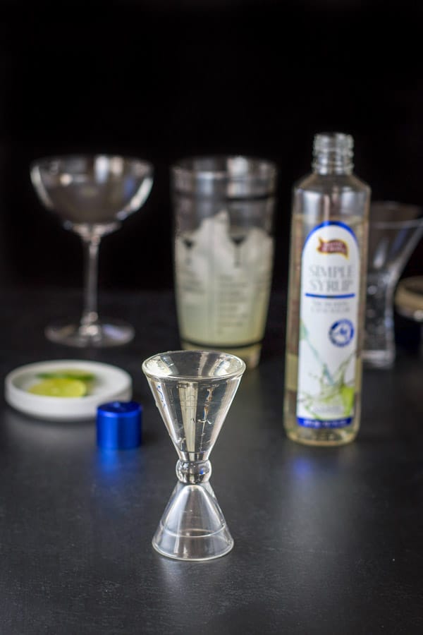 Simple syrup measured out with the bottle, lime garnish half full shaker in the background