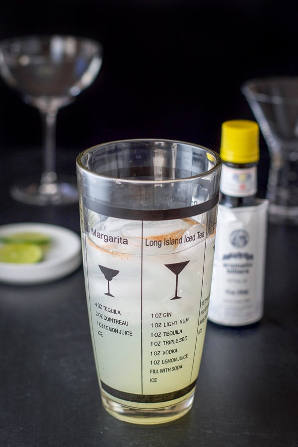 Angostura bitters added to a shaker with the bottle, garnish and glasses behind it