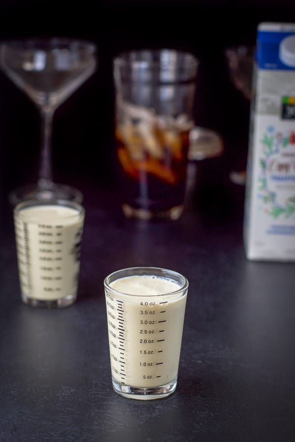 Eggnog poured out along with the carton, filled shaker and glasses in the background