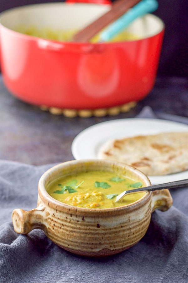 A crock full of lentil soup with naan on a white plate and the red pan in the background