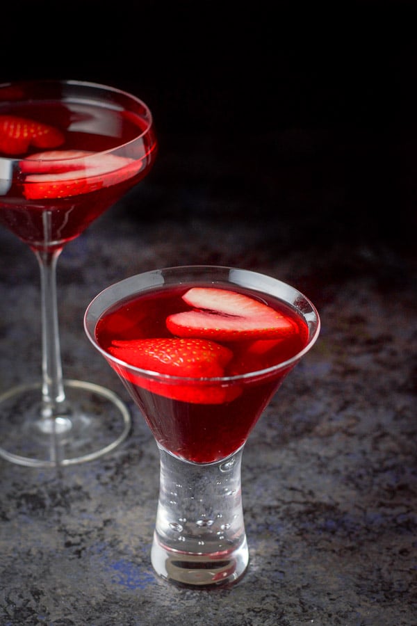 Short glass filled with the martini in front of the other one. There is strawberries floating in the drink