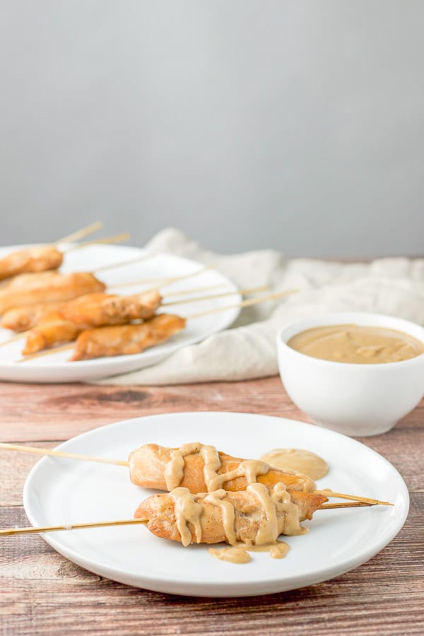 A white plate of 2 chicken skewers with peanut sauce drizzled on it. There is peanut sauce and more skewers in the background