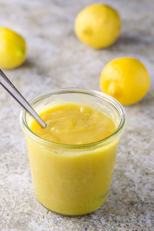 A spoon in the glass jar of lemon curd and whole lemons in the background