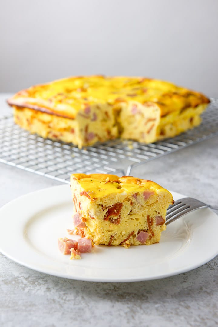 A slice of egg and ham loaf on a salad plate with the whole casserole on a cooling rack behind it.