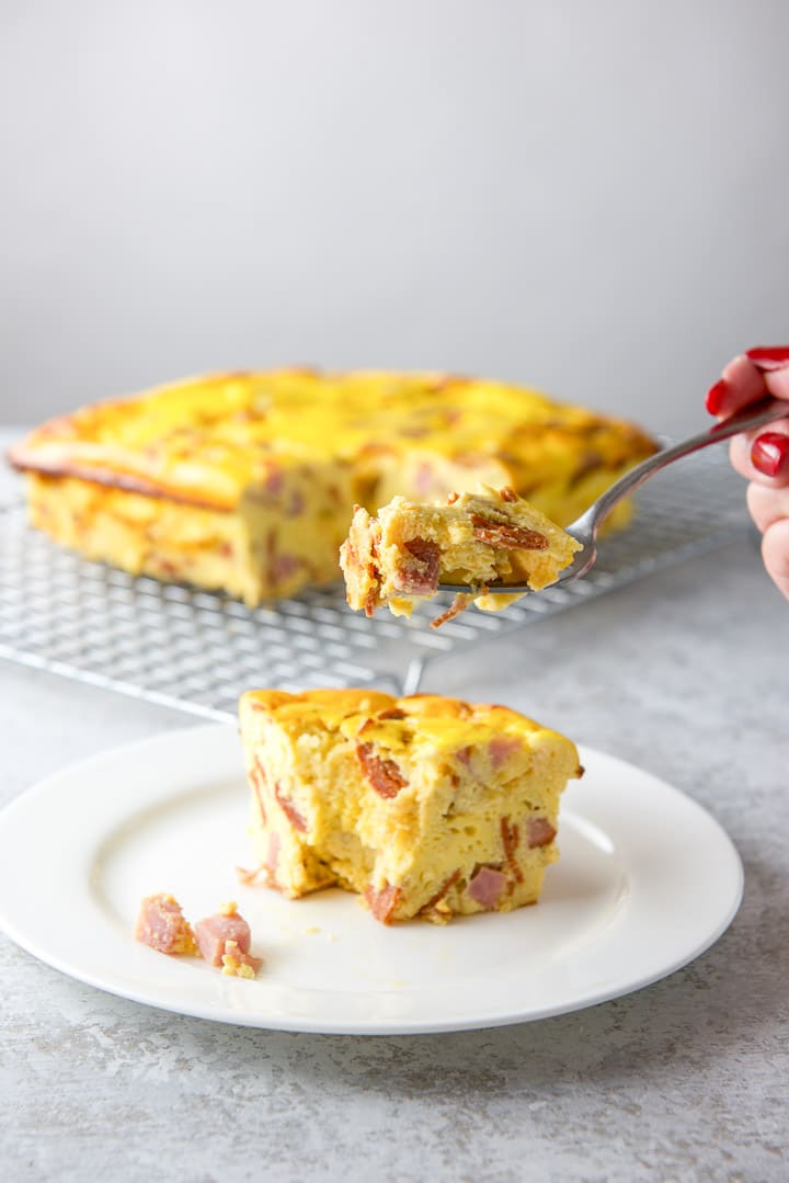 Forkful of egg casserole lifted in the air above the piece on the plate, with the remaining casserole on a cooling rack in the background.