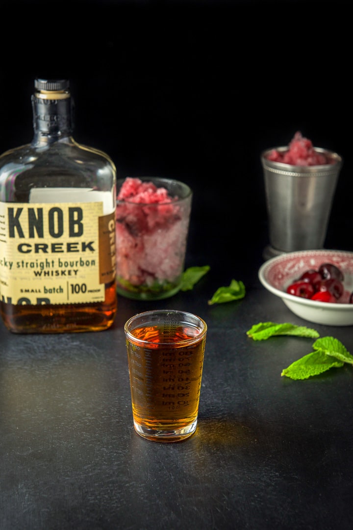 Bourbon measured out with the bottle, cup, glass and some cherries and mint in the background