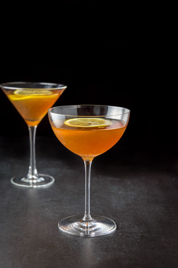 A bowl martini glass filled with the cocktail in front of the classic glass. There are lemon wheels in the drink as garnish