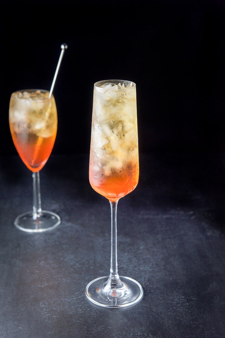 Tall glass in front filled with the orange clear cocktail with another glass with a stirrer in it