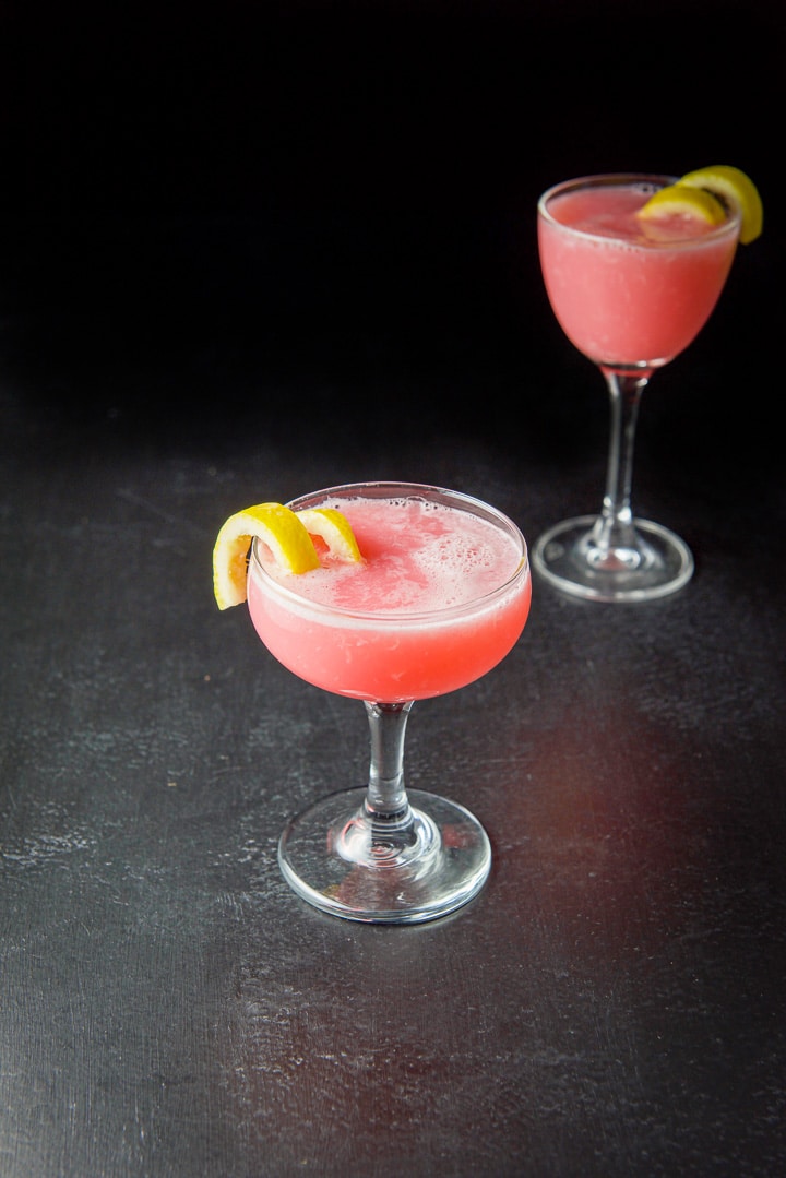 The pink cocktail in a coupe and martini glass with lemon twists as garnish