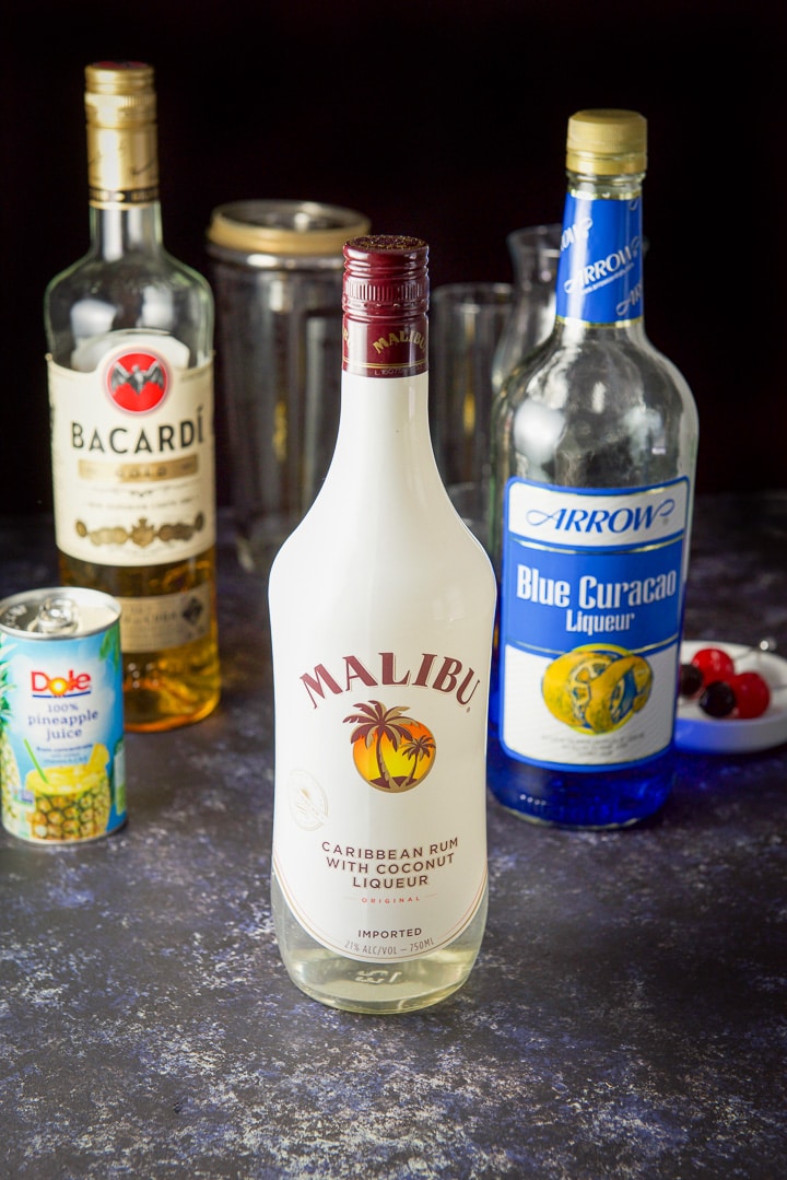 Coconut rum, gold rum, blue curacao and pineapple juice on a table with the garnish, shaker and glassware