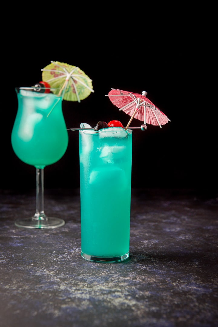 Vertical view of the thin glass filled with the blue cocktail with garnish and tulip glass in the background
