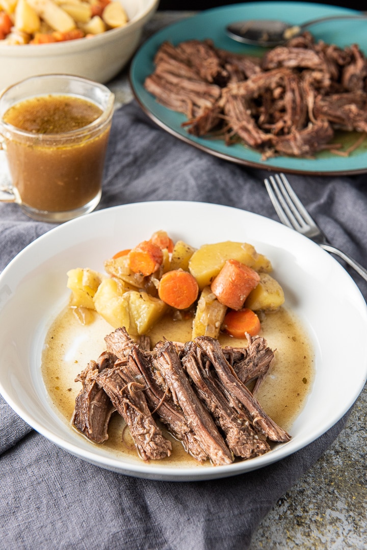 The shredded round roast on a plate with some veggies. There are also veggies in the background as well as gravy and more beef