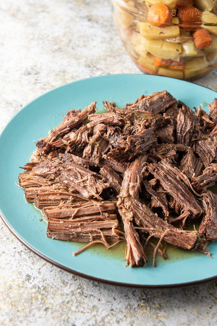 The roast shredded on a blue platter with cooked veggies in the background