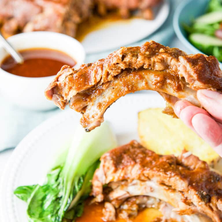 A hand holding one of the baby back ribs over the plate with sauce and more ribs in the background- square