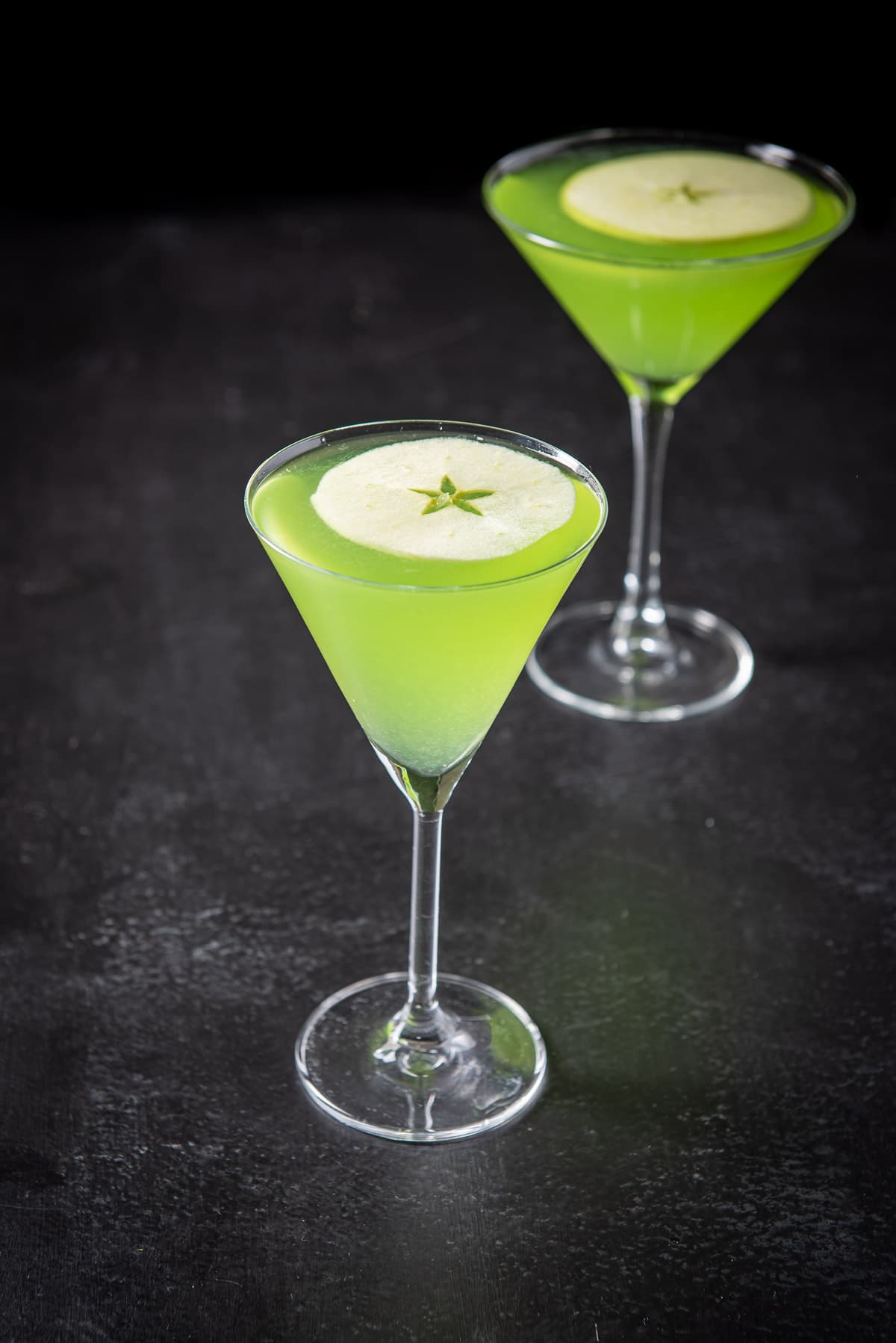 Thin martini glass filled with the apple drink in front of the martini glass behind that