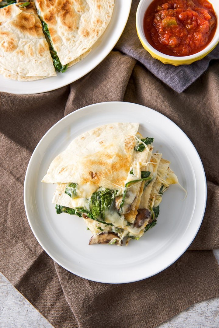 Vertical photo of a stack of quesadillas on a white plate with salsa in the back