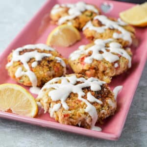 Pink rectangle plate with five crab cakes on it and drizzled with sauce - square