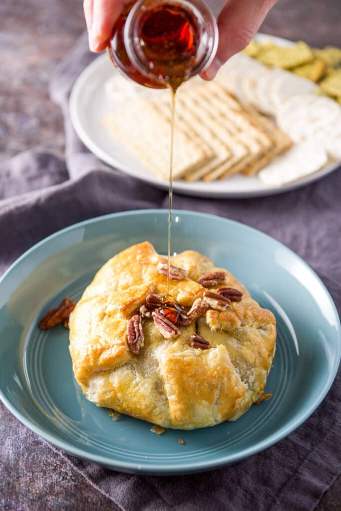Pecans and maple syrup being poured on the brie with crackers in the background