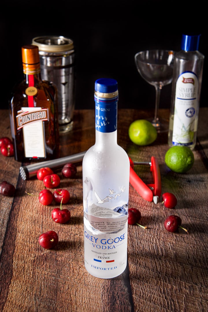 Vodka, limes, Cointreau and simple syrup on the wooden table. There are also a bunch of cherries, muddler and cherry pitter on the table too