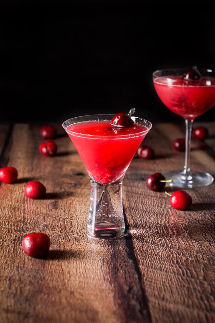 The bubble martin glass filled with the cherry cosmo with the bowl glass behind it. There are lots of cherries strewn on the table