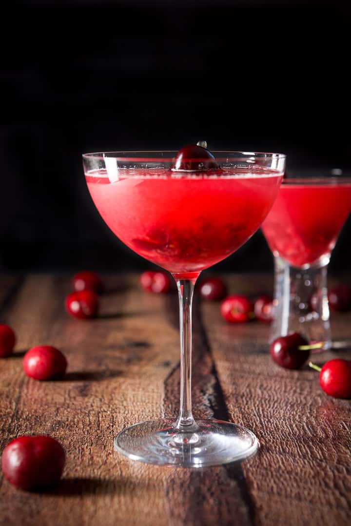 Vertical view of the bowl glass filled with the cosmo with the bubble glass behind it and there are cherries on the table