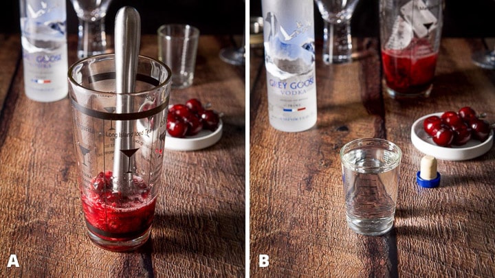 Muddled cherries in the cocktail shaker with a muddler in there as well. Vodka poured out with the vodka bottle in the background as well as the cherry garnish on picks