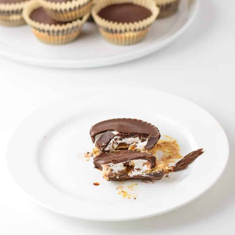 A chocolate marshmallow cup bitten into on a white plate. There are more chocolate cups behind it on a plate
