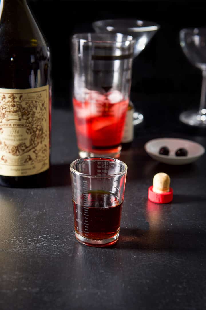 Sweet vermouth poured out with the bottle, half filled shaker, glasses and cherries in the background