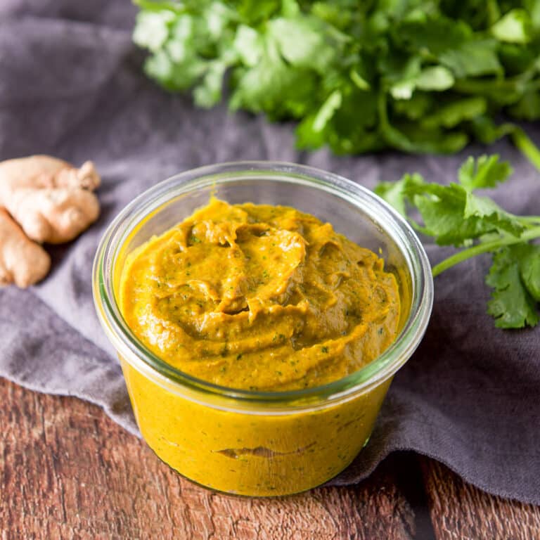 A short glass jar filled with curry paste with cilantro and ginger in the background