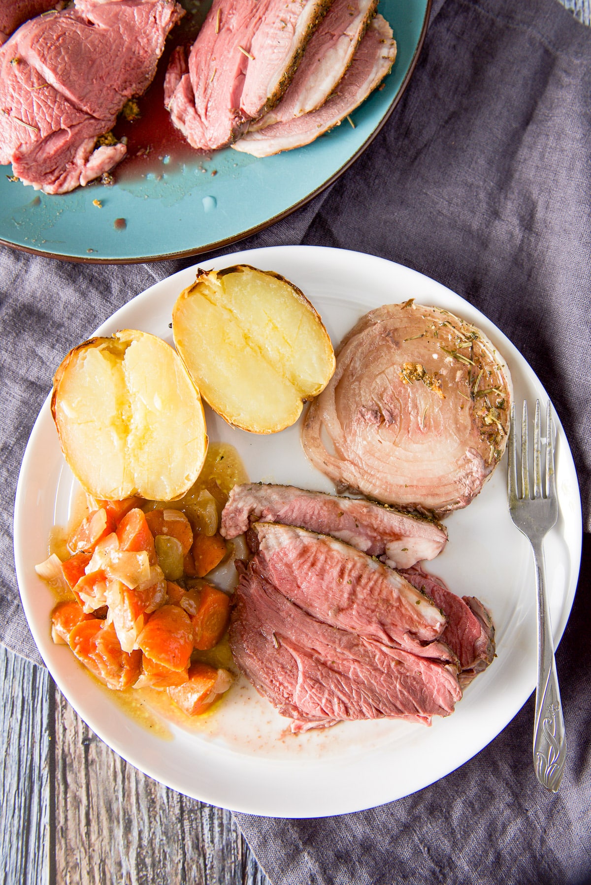Overhead view of a white plate with a few slices of lamb, carrots, baked potato and onion