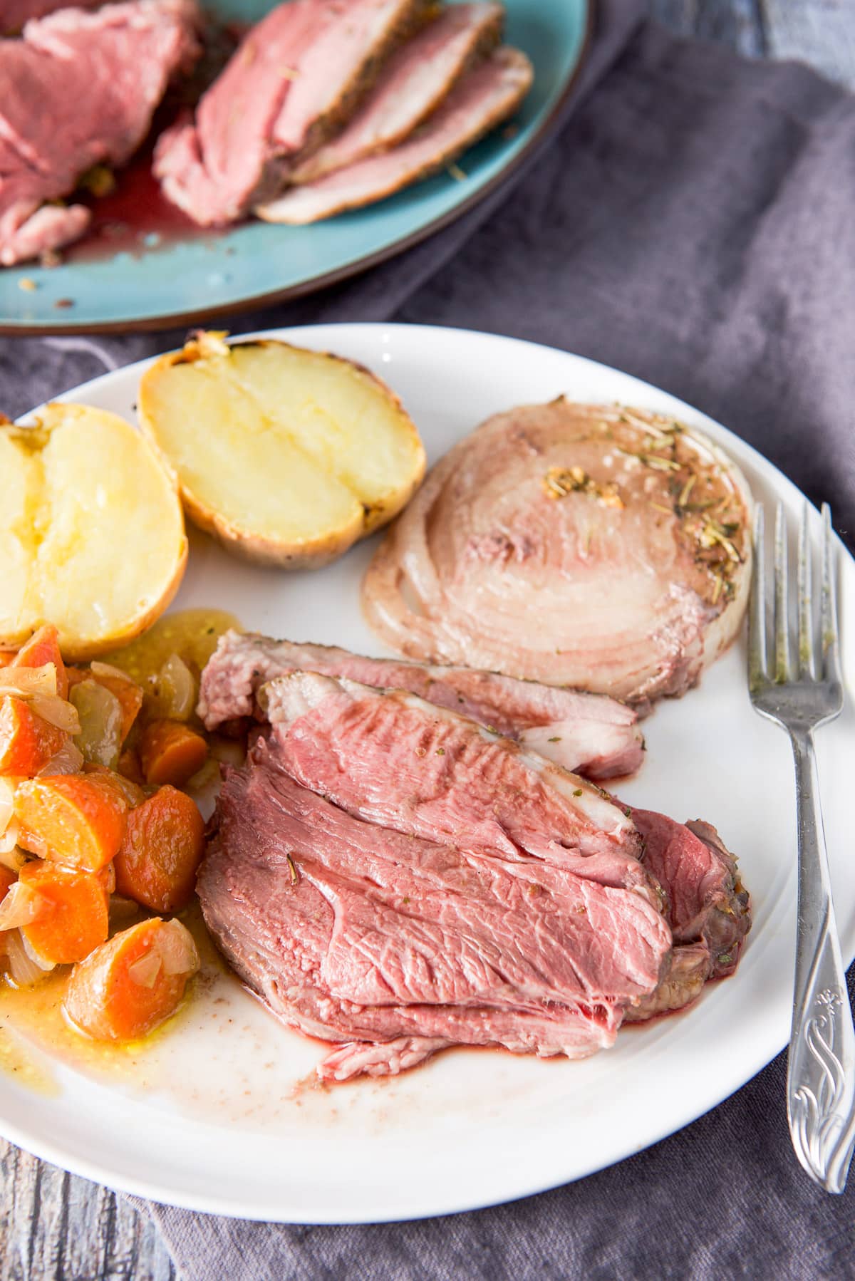 A white round plate with sliced lamb, onion, potato and carrots with the platter of the lamb in the background