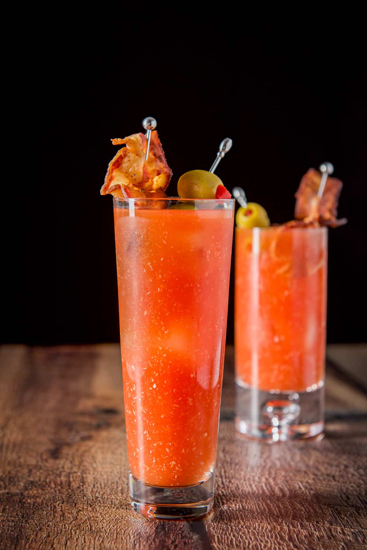 Vertical view of the flared glass in front of the high ball glass filled with the cocktail and olives and bacon as garnish