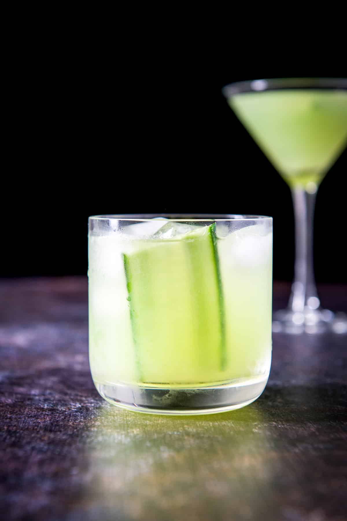The cucumber gimlet in a double old fashioned glass with the classic martini in the back. There is a cucumber slice in it as garnish