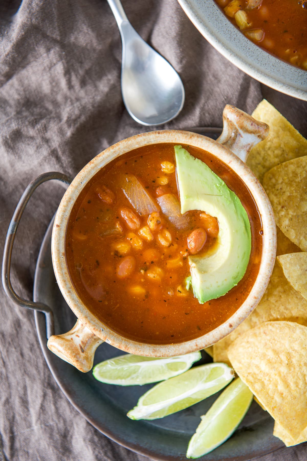 Overhead view of the soup in a crock with avocado, lime wedges and chips