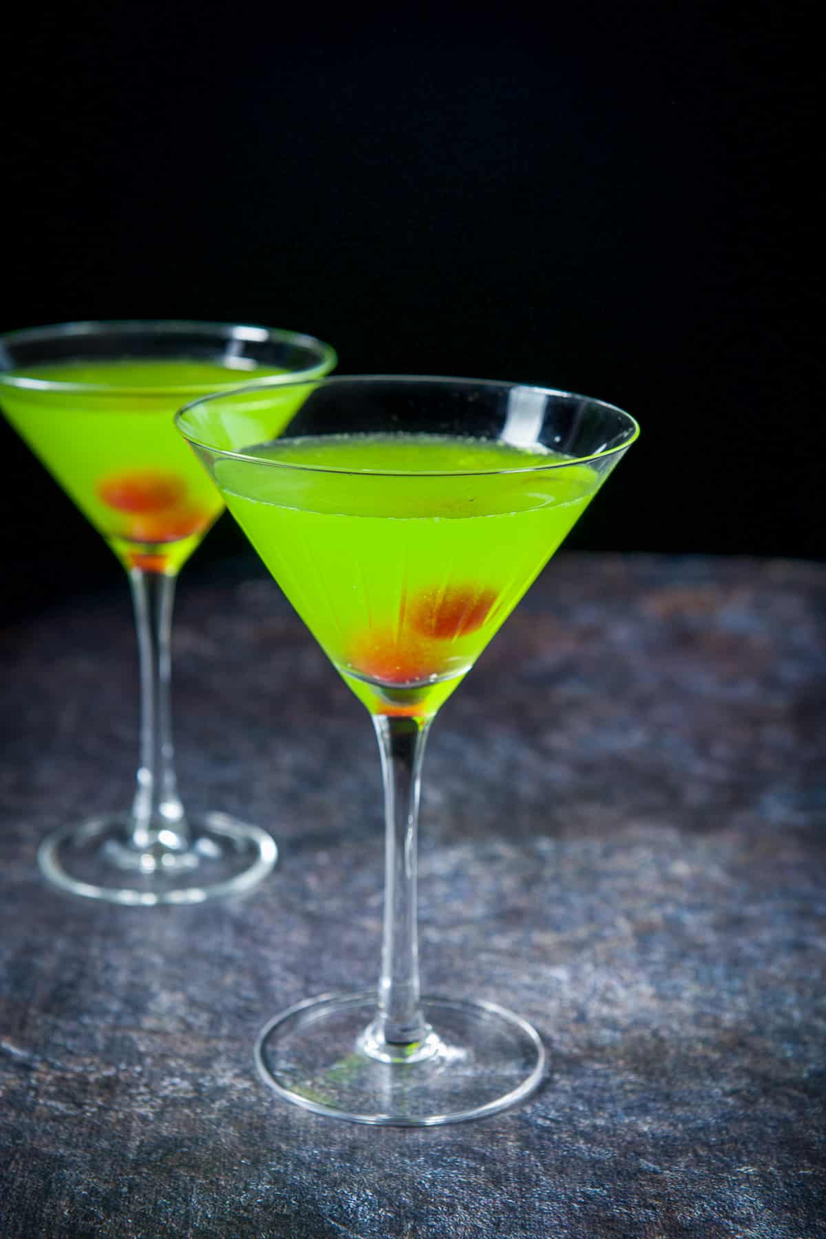 Two martini glasses filled with the green drink with cherries in the glasses as garnish