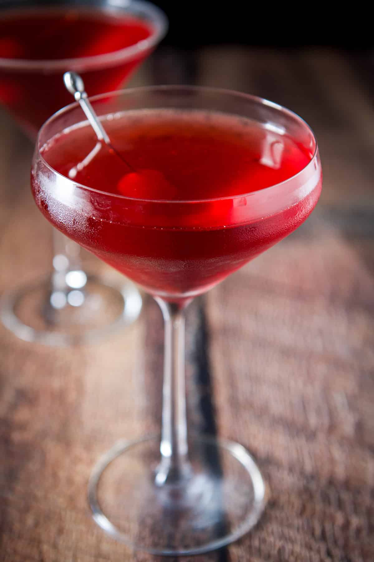 Close up of the curved glass filled with the red cocktail and a cherry and pick in it