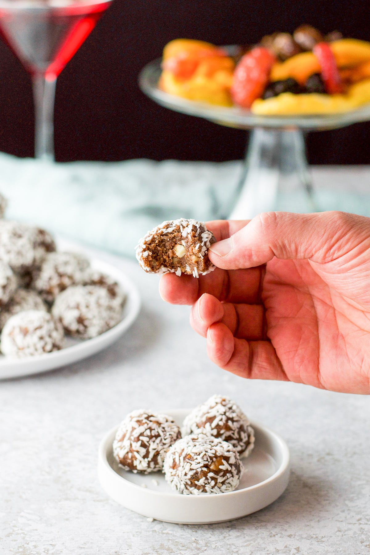 A hand holding a coconut covered ball with a bite out of it with dried fruit in the background