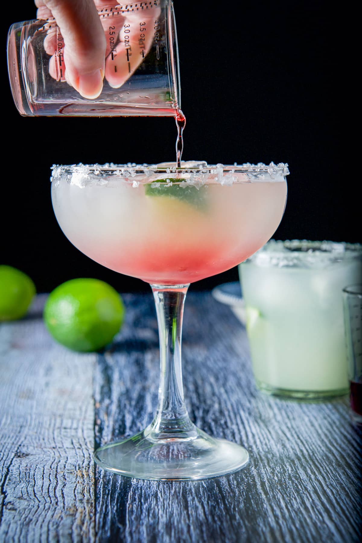 Chambord being poured into the margarita glass with a glass and limes in the back