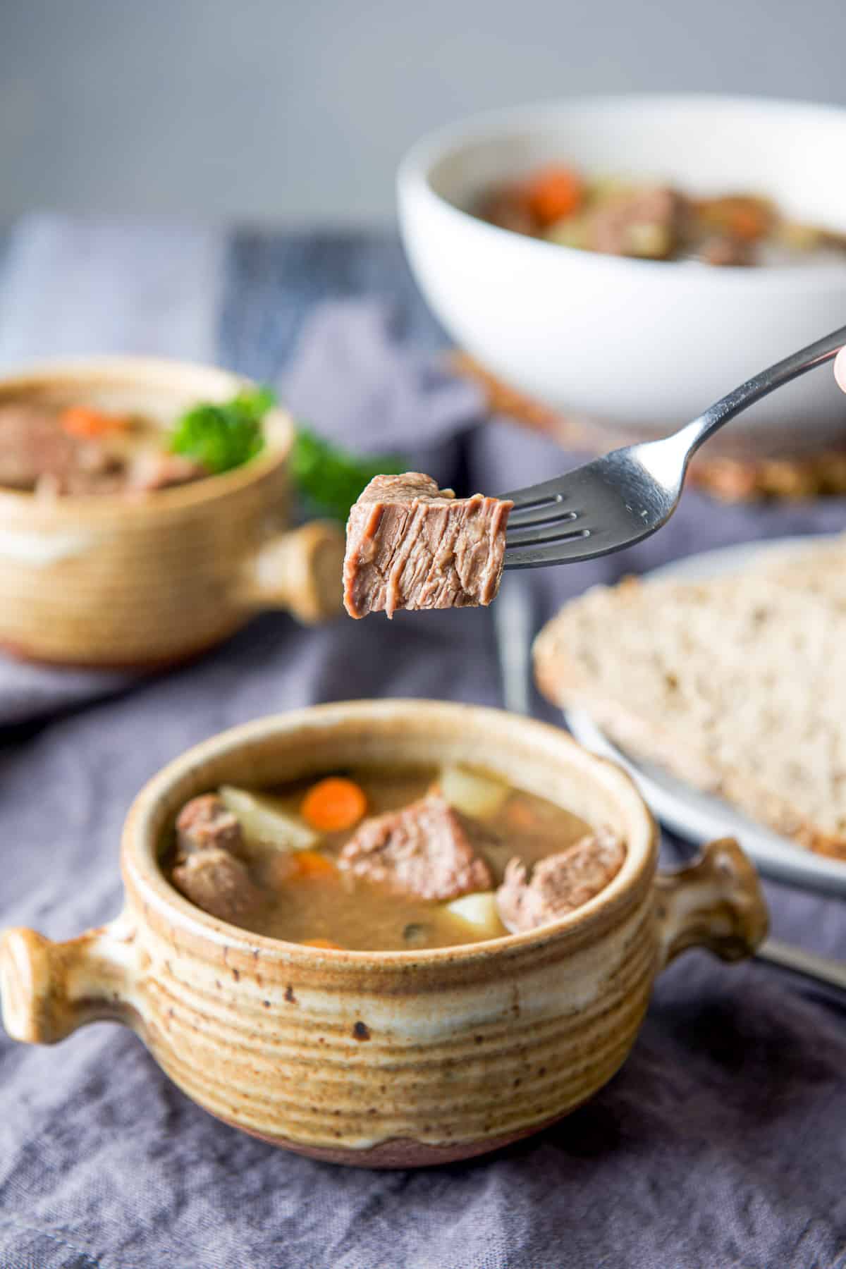 A fork holding a piece of lamb over the soup crock with another crock, bread and white bowl in the back