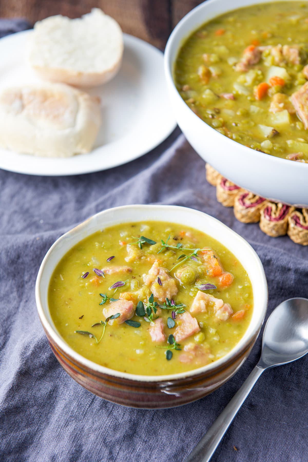 A brown bowl with pea soup, a big white bowl in back and a roll