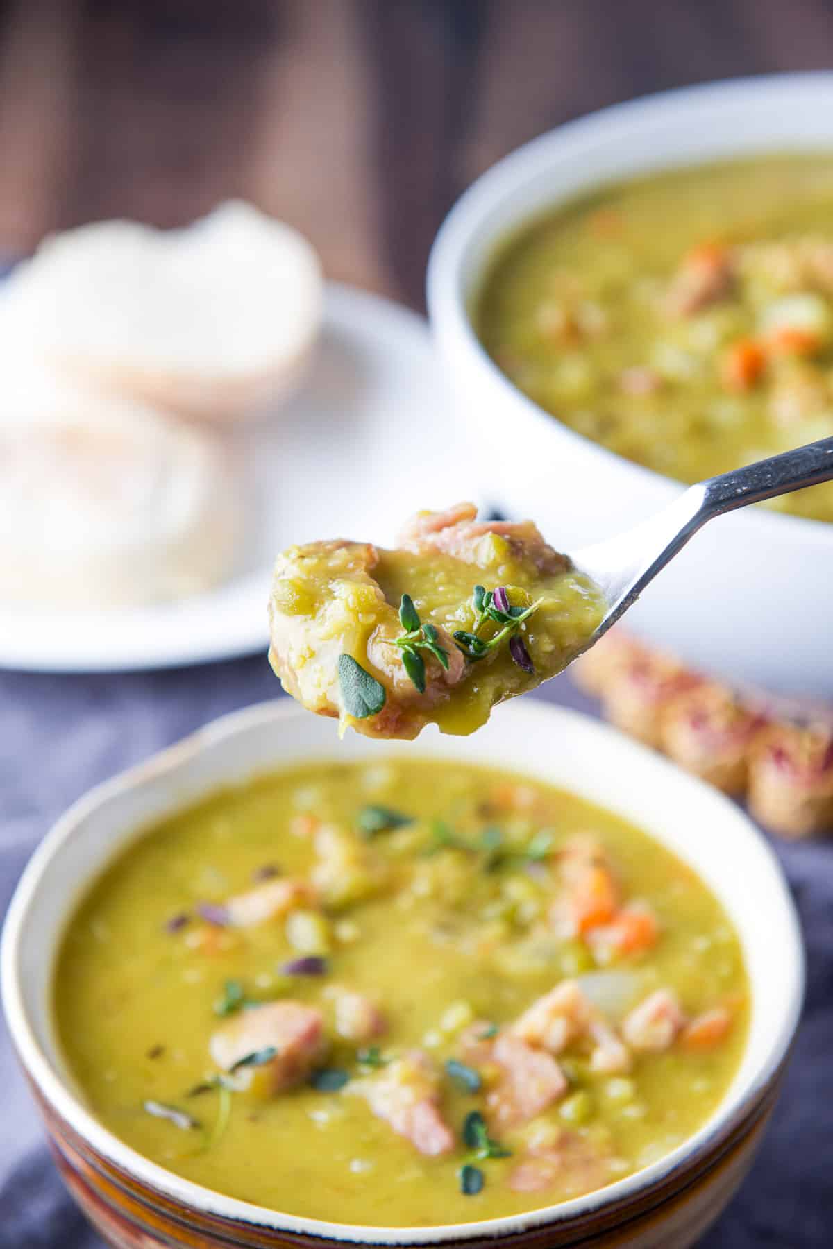 A spoon with soup in it held over a bowl of soup, and big bowl and roll in the back